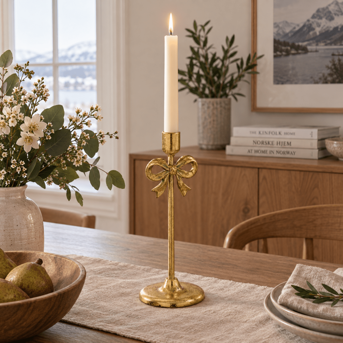 Gullfarget lysestake med dekorativ sløyfe og tent kronelys, plassert på et spisebord i et lyst nordisk hjem.