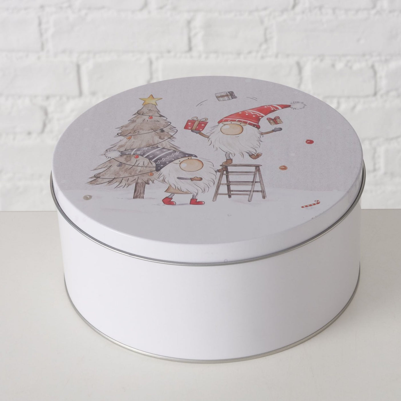 Hvit kakeboks med julemotiv av to nisser som pynter tre, én på stige, mot lys mursteinsvegg.