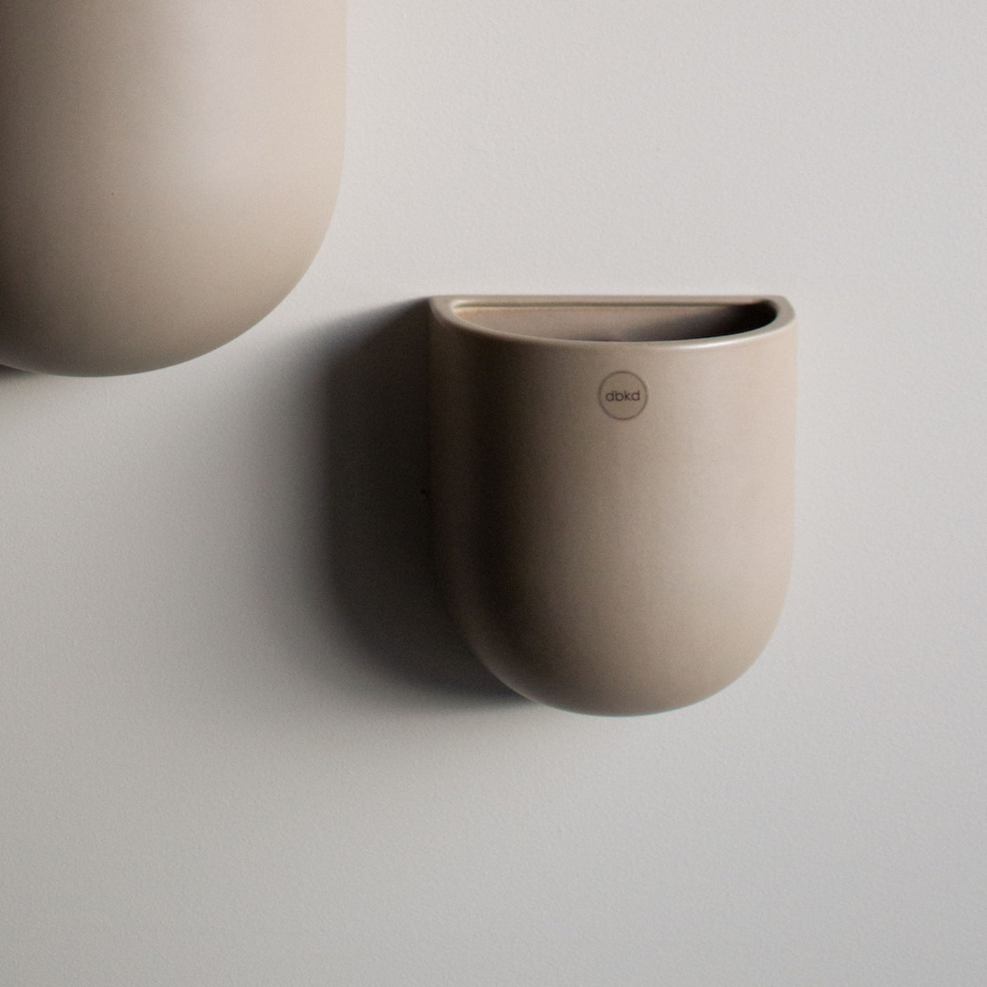 Lys keramikkpotte fra dbkd henger på en lys vegg, delvis i ramme med en større potte.