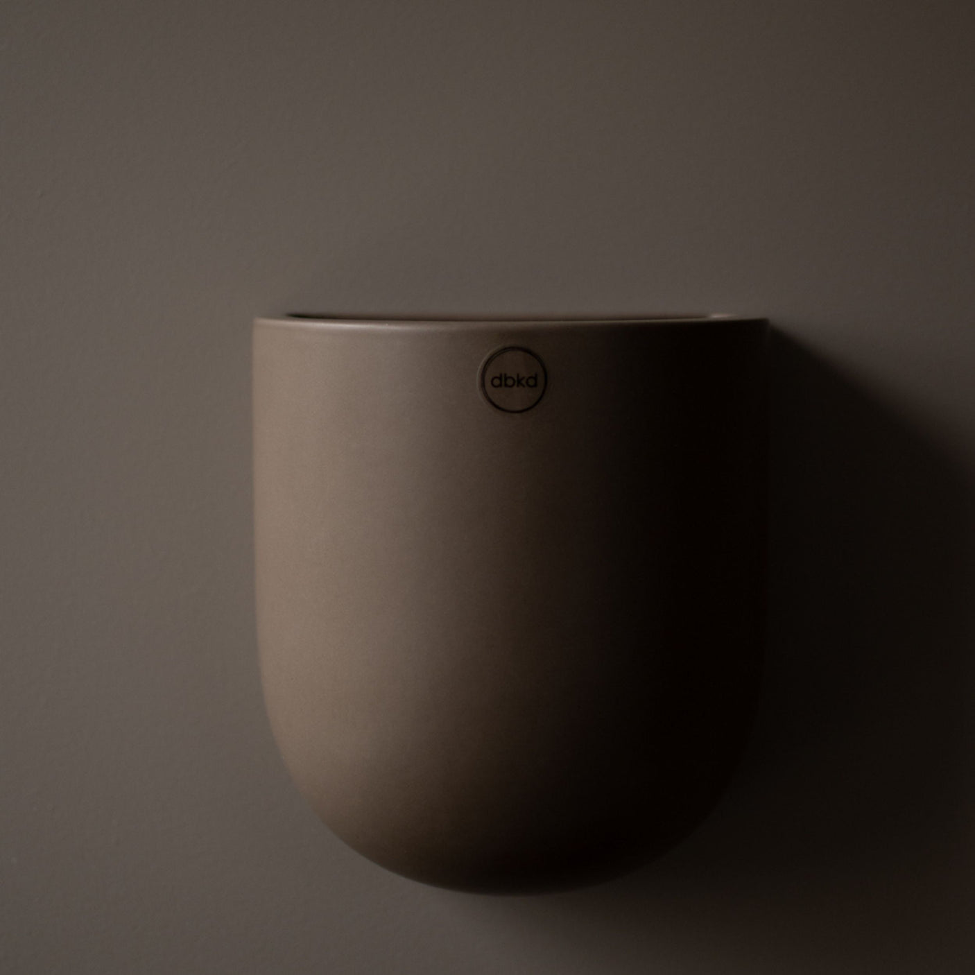 Mørk keramikkpotte fra dbkd henger alene på vegg med skyggefall.
