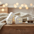 Tre keramikkhus med tent lys i vinduene, plassert koselig på bord i stue med dempet belysning.
