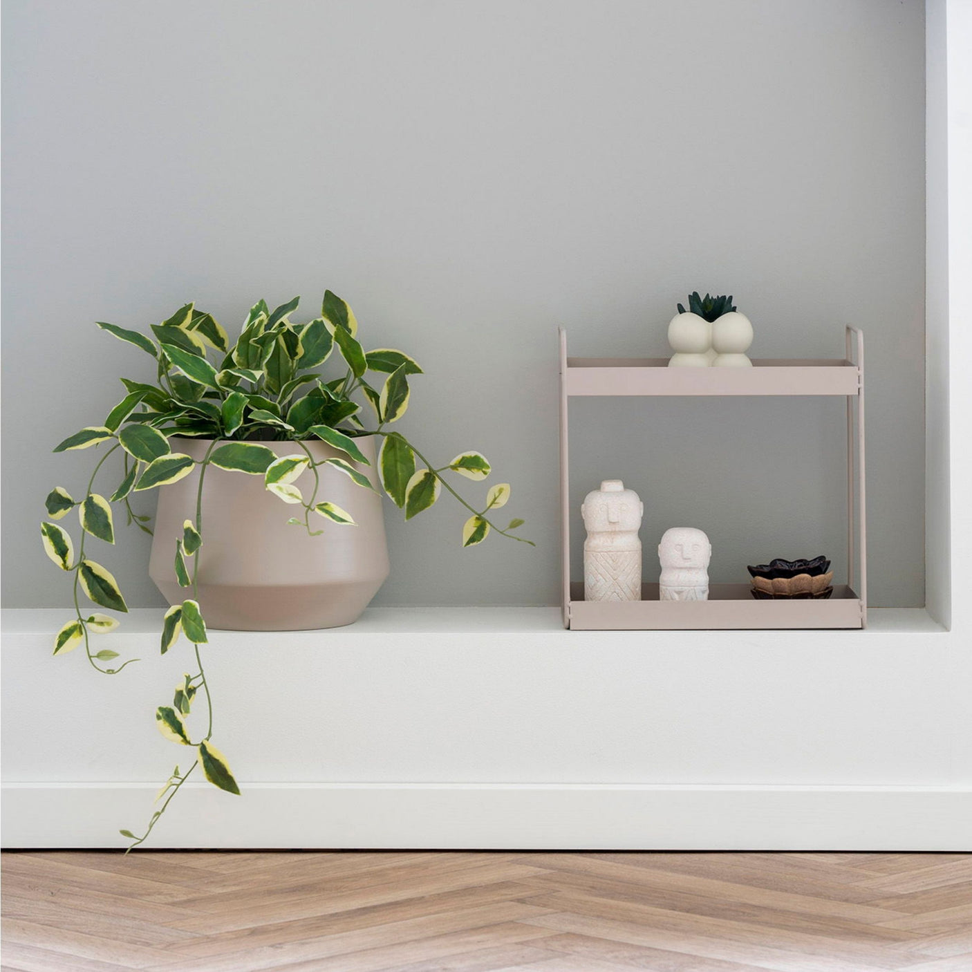 Beige metallhylle plassert i veggnisje med dekorfigurer og planter, ved siden av grønn potteplante.