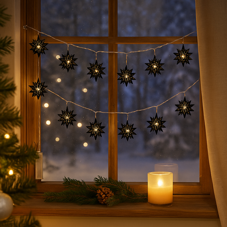 LED-lyslenke med sorte stjerner i tre henger i et vindu, lyser varmt og skaper julestemning med snødekt landskap utenfor og granbar på vinduskarmen.