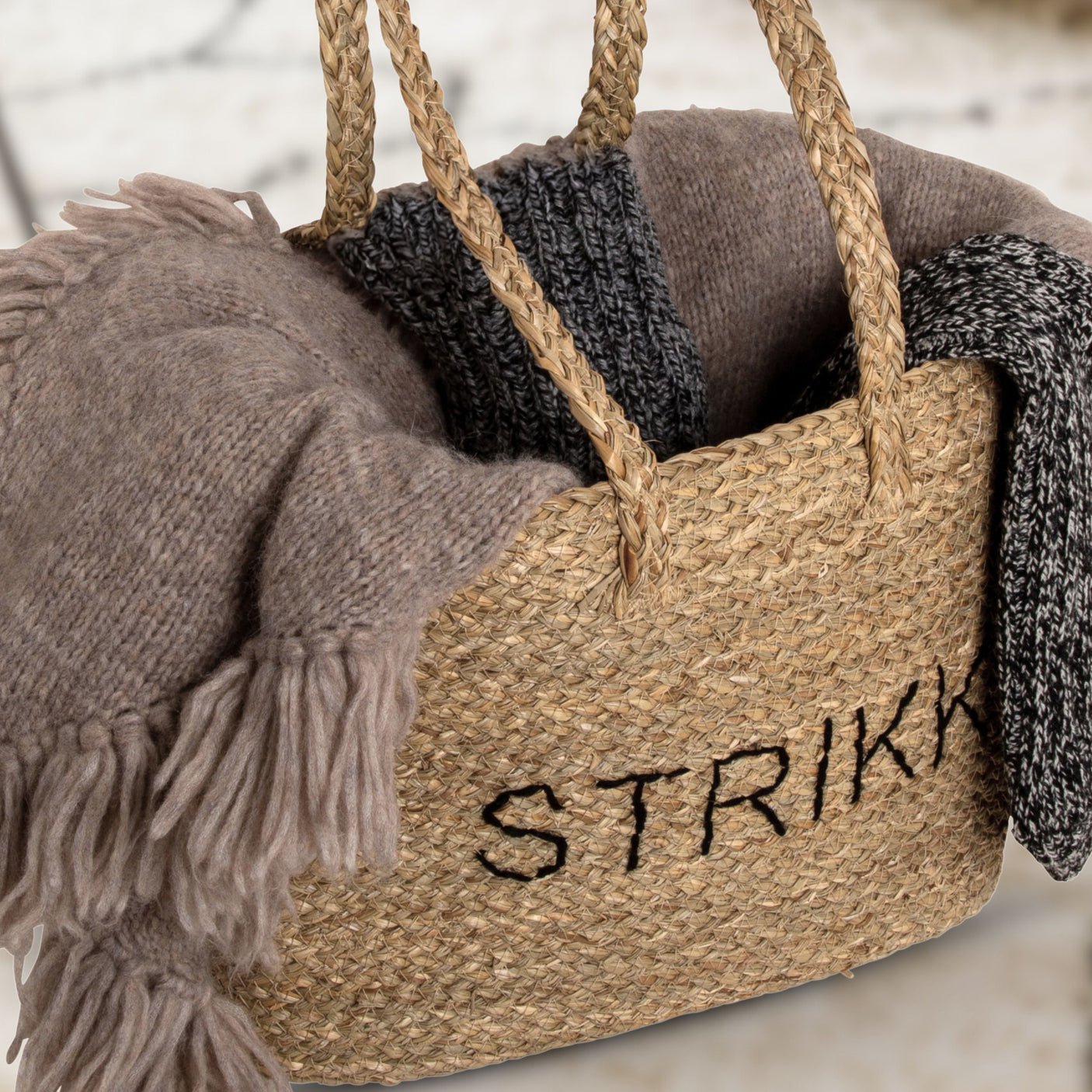 Stråveske med teksten STRIKK fylt med ullgensere og strikkeprosjekter i varme, nøytrale toner.
