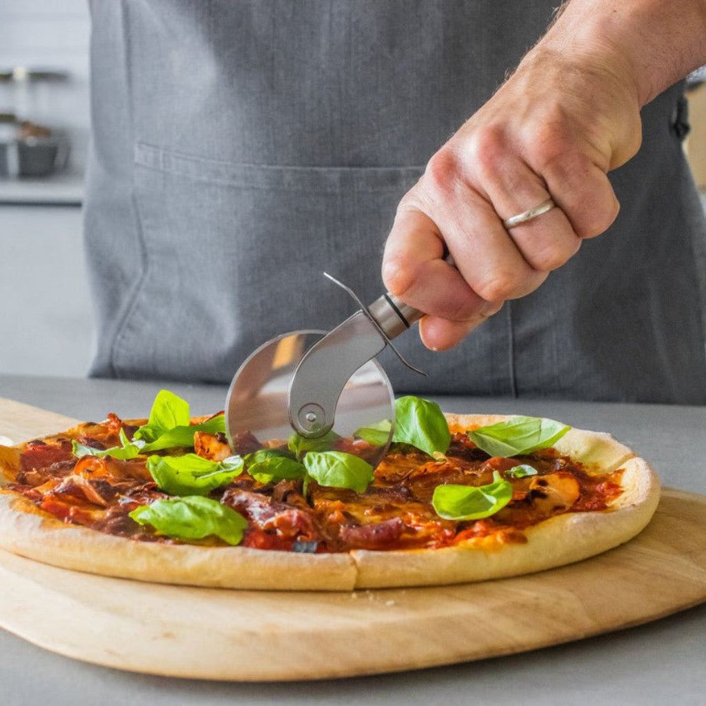 Hånd skjærer en hjemmelaget pizza med pizzahjul i rustfritt stål på en rund trefjøl. - Lindas dekor