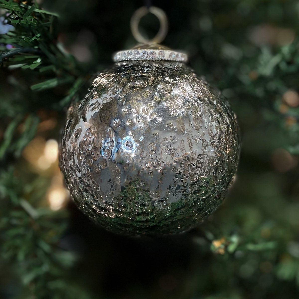 Grå julekugle i glas – hængende på juletreet med grønne greiner og julelys i baggrunden.