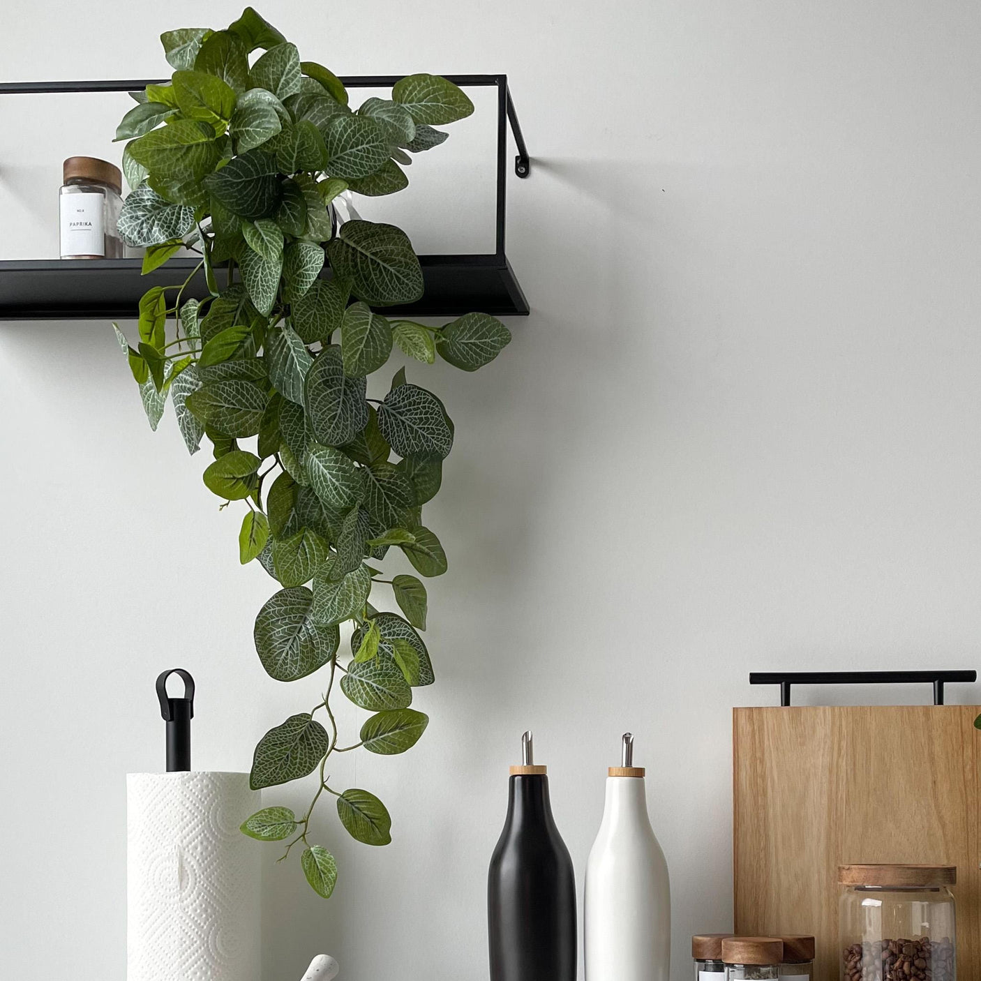 Kunstig Fittonia-plante på sort hylle på kjøkken, med tilbehør på benken under.
