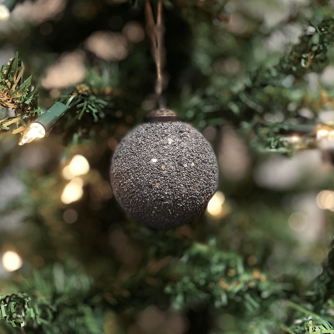 Mørk grå julekule med glittereffekt henger på grønt juletre med små varmhvite julelys.
