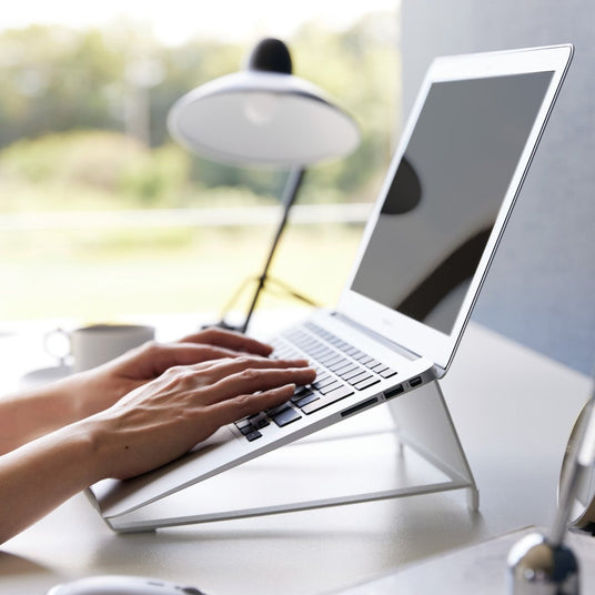 Ergonomisk laptopstativ fra Yamazaki i hvit stål – person skriver på MacBook løftet til øyehøyde, skrivebordslampe og vindu i bakgrunnen