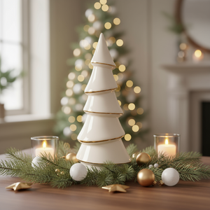 Kremfarget keramikkjuletre med gullkant på dekorfat med røde kuler og tente lys foran juletre og peis.