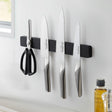 Sort magnetisk knivholder montert på kjøkkenvegg med kniver og saks.