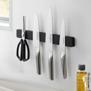 Sort magnetisk knivholder montert på kjøkkenvegg med kniver og saks.