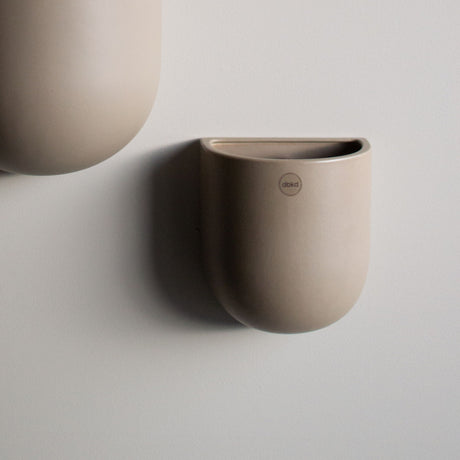 Lys keramikkpotte fra dbkd henger på en lys vegg, delvis i ramme med en større potte.