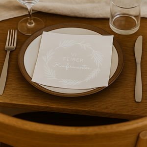 Serviett med teksten «Vi feirer konfirmanten» ligger på tallerken på et dekket trebord i naturlig lys.
