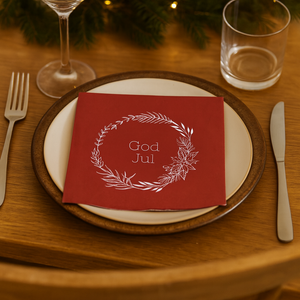 Rød serviett med teksten «God Jul» ligger på tallerken omgitt av julegrønt og dempet lys.
