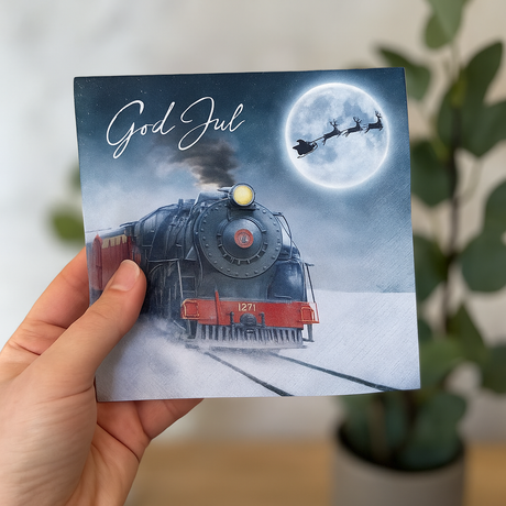 Serviett God Jul Lokomotiv snøfrost – juleserviett med tog og måne motiv, holdt i hånd foran nøytral bakgrunn med grønne planter. Perfekt som julepynt og til dekking av julebord.