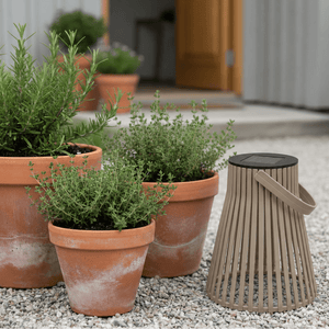 Brun solarlykt plassert på grus ved et inngangsparti, omgitt av terrakottapotter med grønne urter og planter. - Lindas dekor 