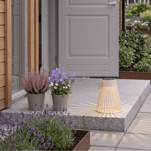 Beige solarlykt med varmt lys plassert ved inngangspartiet til et hus, omgitt av blomsterpotter og planter. - Lindas dekor 