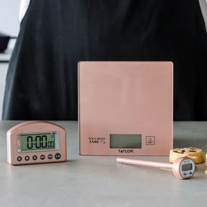 Kjøkkenvekt, timer og steketermometer i rosé i bruk under baking på kjøkkenbenken